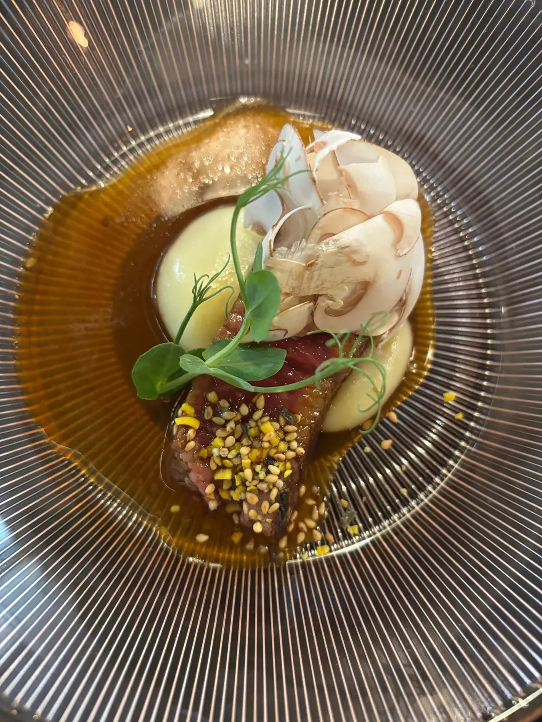 A plated dish at Kuvo in a ribbed glass bowl, with sliced mushroom, pea shoots and a golden broth