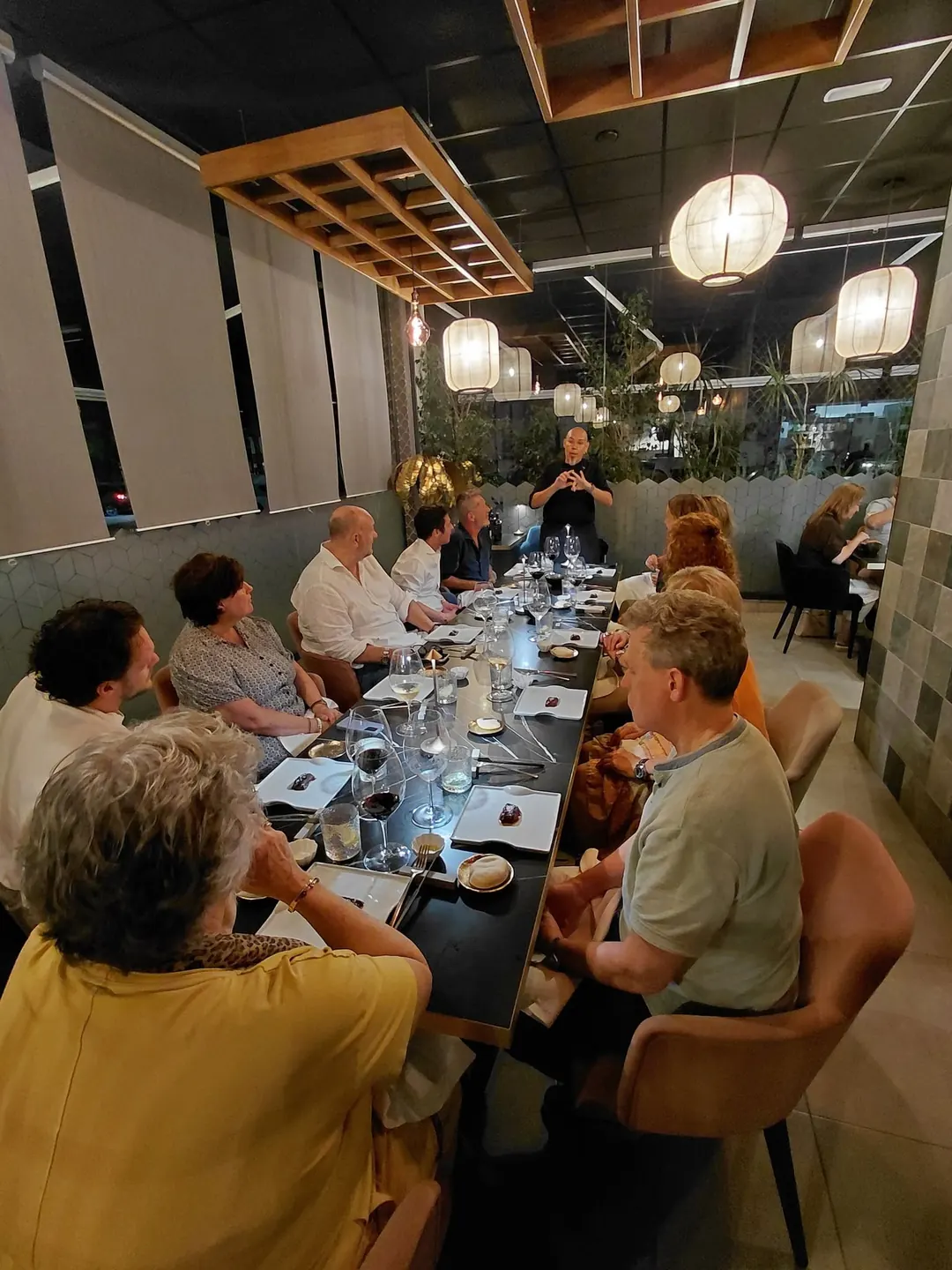 Guests around a long table at dinner, with Óscar talking to them at the far end