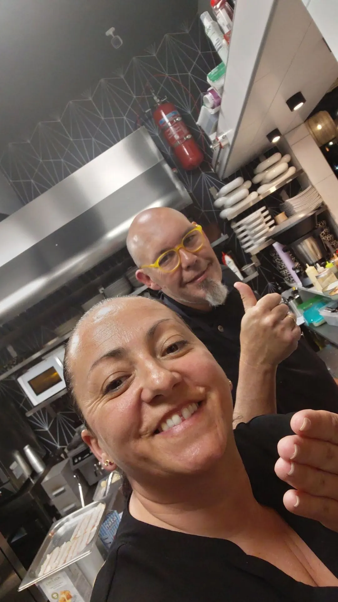 Pilar smiling at the camera in the kitchen, with Óscar giving a thumbs up behind her